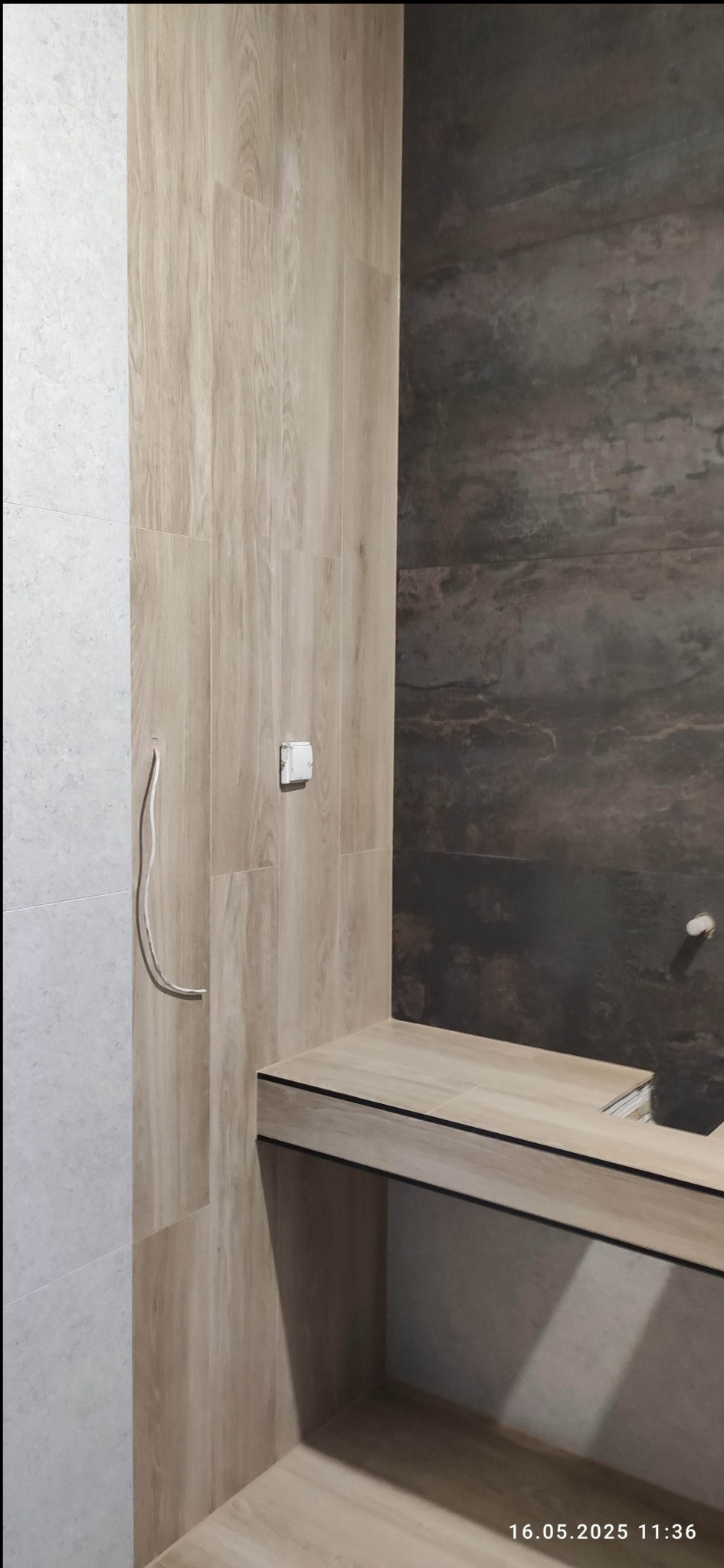 Wykończona łazienka z płytkami imitującymi drewno i kamień, widoczne gniazdko i wystający kabel. Umywalka w zabudowie z płytek.