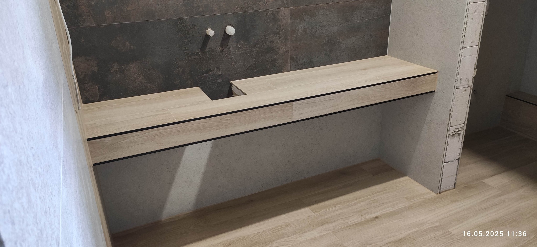 Łazienka w trakcie wykończenia: drewnopodobna zabudowa pod umywalkę z otworem, szara płytka na ścianie, widoczne instalacje wodne, podłoga drewnopodobna.