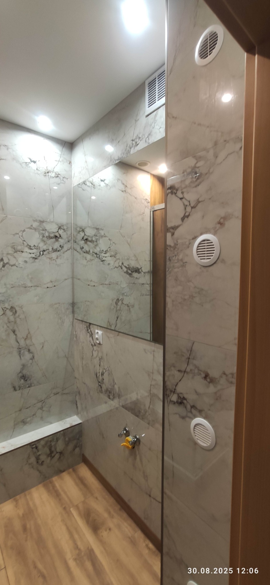 Wykończona łazienka z marmurowymi płytkami na ścianach i drewnopodobną podłogą. Widoczne detale: kratki wentylacyjne, bateria umywalkowa i lustro. Nowoczesny design.