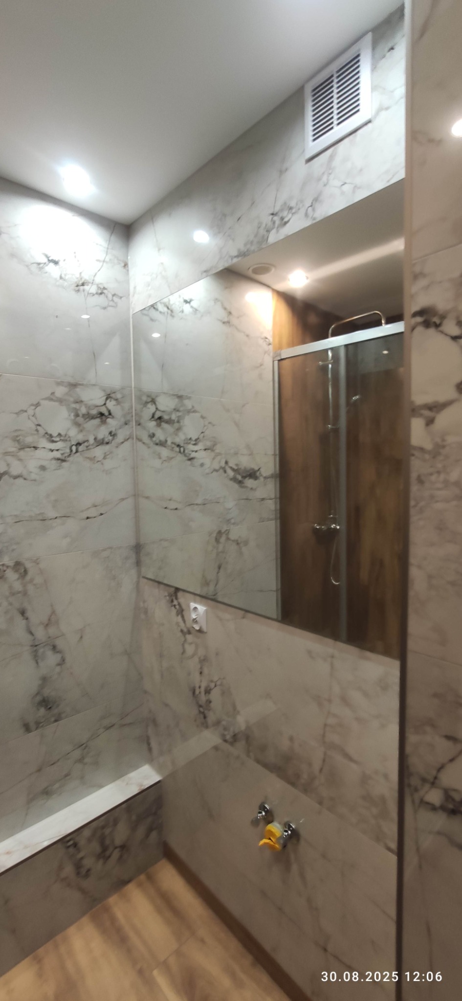 Wykończona łazienka z marmurowymi płytkami na ścianach i podłodze. W lustrze odbija się kabina prysznicowa z drewnianymi panelami. Widoczna kratka wentylacyjna.
