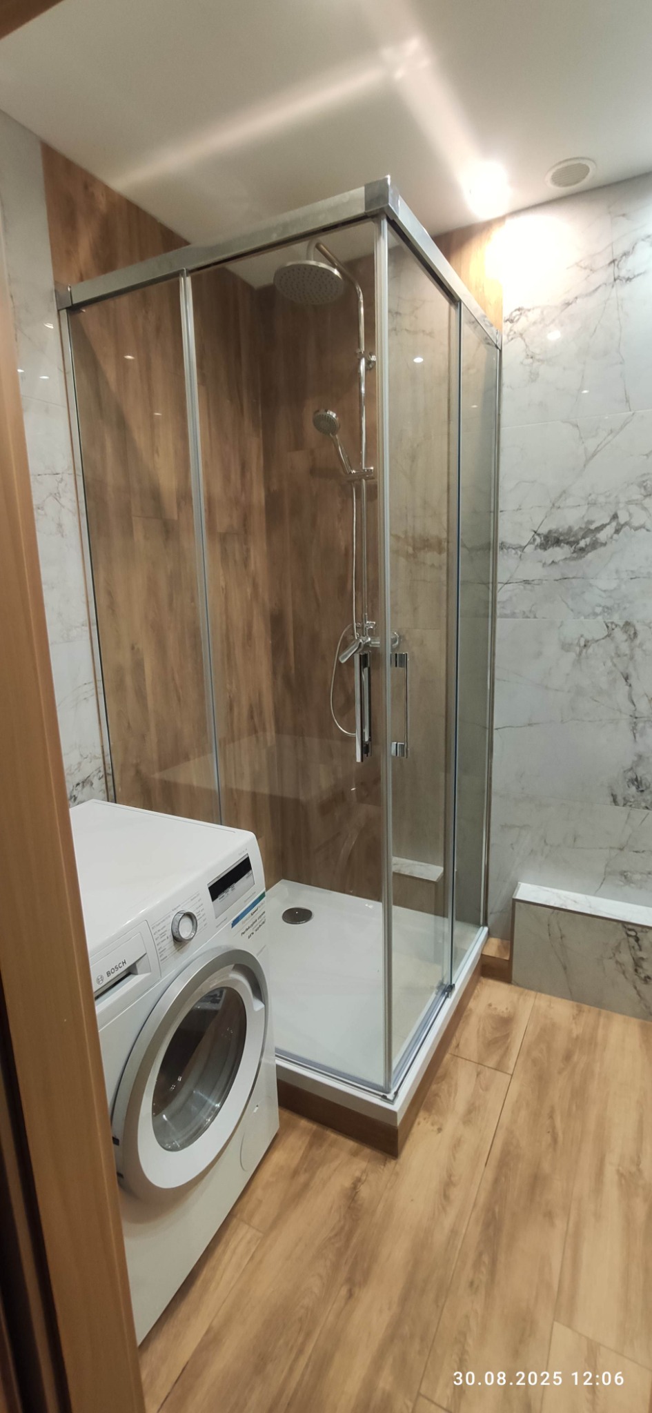 Nowoczesna kabina prysznicowa z deszczownicą w wykończonej łazience, obok pralka Bosch. Drewnopodobna i marmurowa okładzina ścienna, podłoga z paneli.