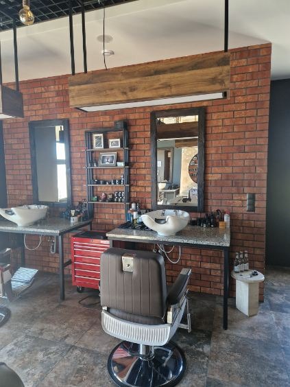 Nowoczesne wnętrze salonu fryzjerskiego z ceglaną ścianą, dwoma stanowiskami z umywalkami i fotelami. Widoczna półka z kosmetykami i narzędziami.