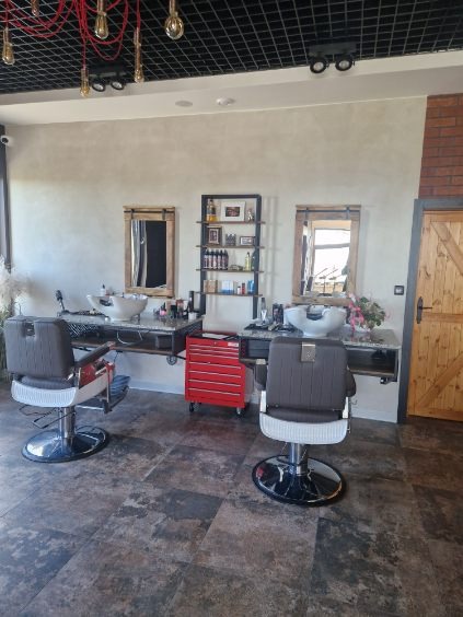 Nowoczesne wnętrze salonu fryzjerskiego z dwoma stanowiskami, lustrami w drewnianych ramach i dekoracyjnym oświetleniem. Czerwony wózek narzędziowy dodaje koloru.