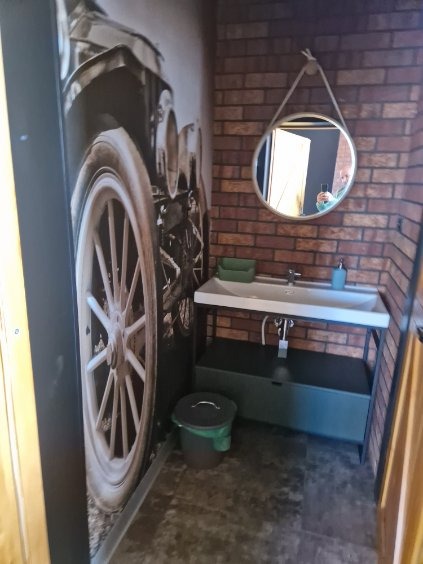 Łazienka w stylu retro z tapetą z motywem koła samochodu, ceglaną ścianą i okrągłym lustrem na sznurku. Umywalka na metalowym stelażu, kosz na śmieci.