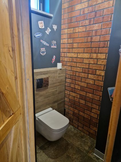 Wykończona toaleta w stylu loft z ceglaną ścianą i drewnianymi drzwiami w Nowym Tomyślu. Na ścianie naklejki. Widok na muszlę klozetową i płytki.