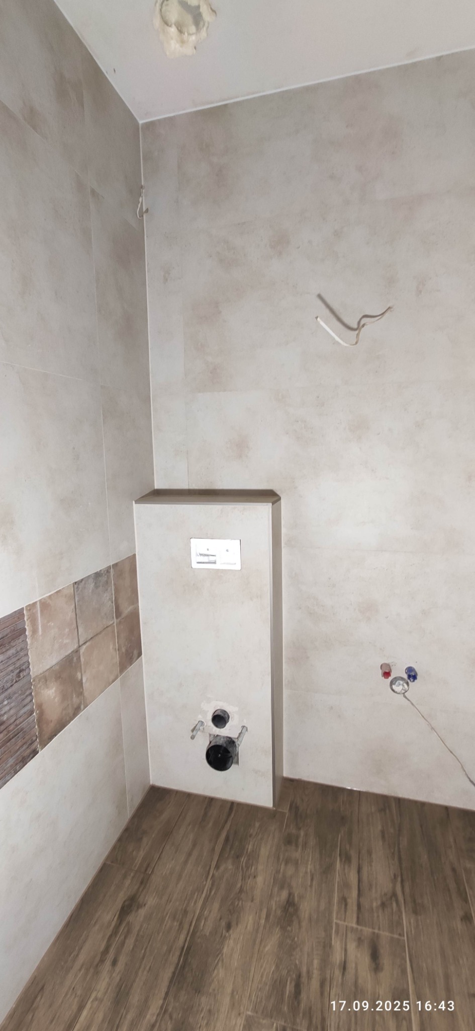 Wnętrze łazienki w trakcie wykańczania: widok na stelaż podtynkowy WC z przyłączami, częściowo ułożone płytki ścienne i podłogowe imitujące drewno.