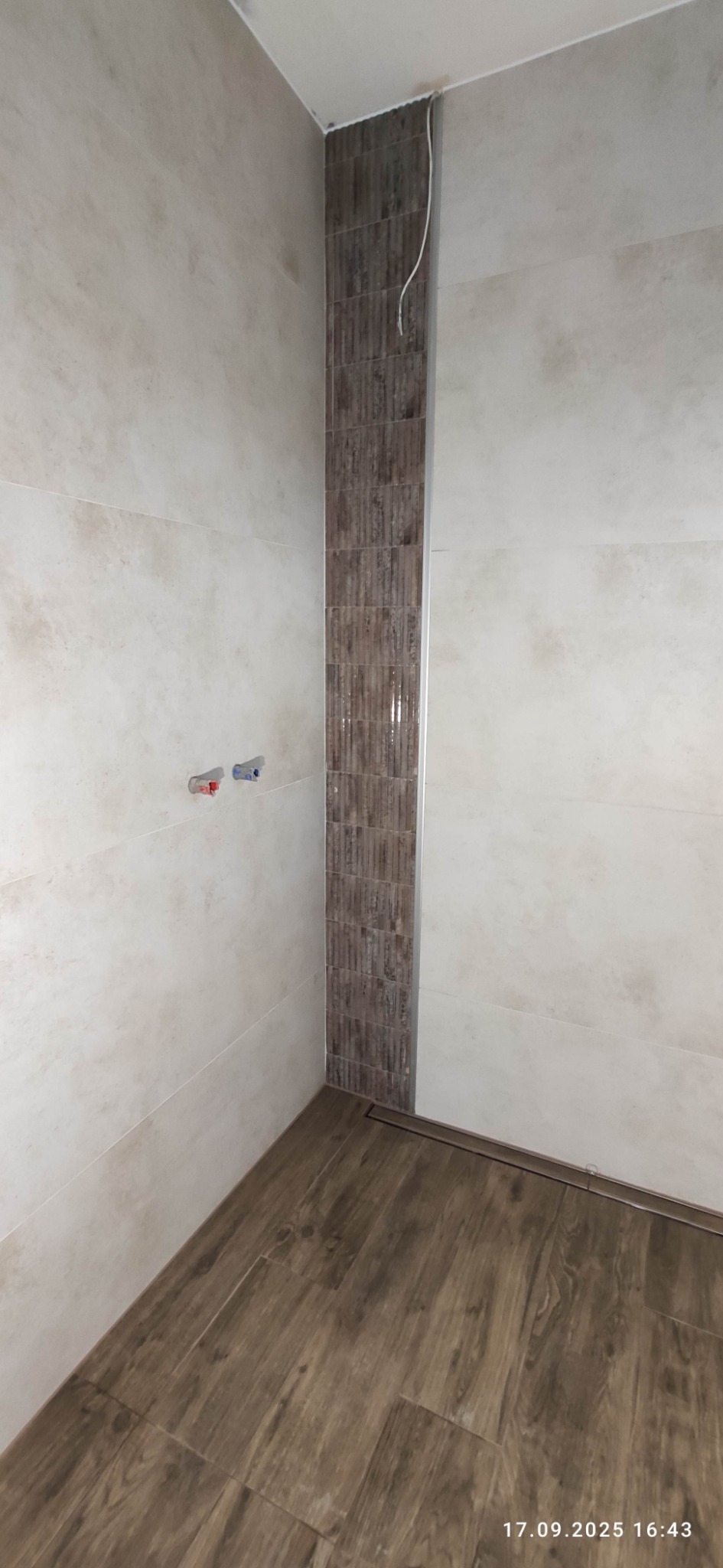 Wykończona łazienka z beżowymi płytkami na ścianach i brązowymi na podłodze. Pionowy pas dekoracyjny z mozaiki. Widoczne przyłącza wodne i odpływ liniowy.