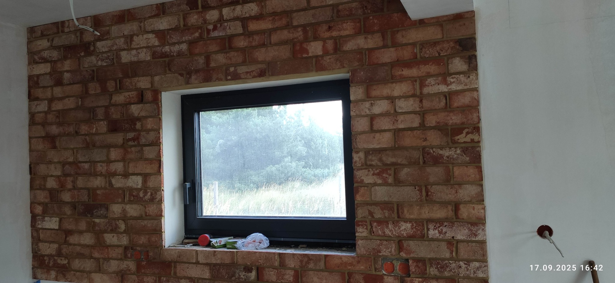Ściana z cegły klinkierowej wokół okna z widokiem na zieleń. Widoczne puszki elektryczne i akcesoria glazurnicze na parapecie okiennym.