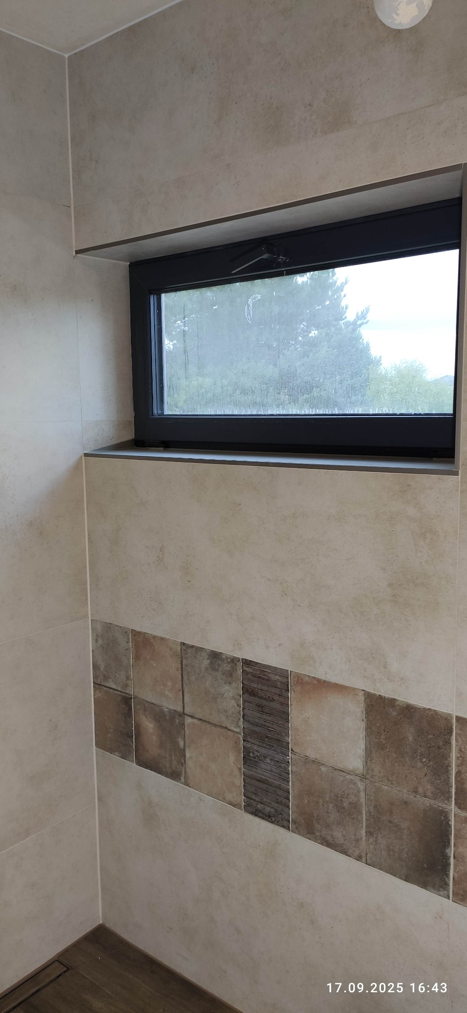 Wykończona łazienka z oknem: beżowe płytki na ścianach, dekoracyjny pas z brązowych płytek, czarna rama okna z widokiem na zieleń. Nowoczesne i estetyczne rozwiązanie.