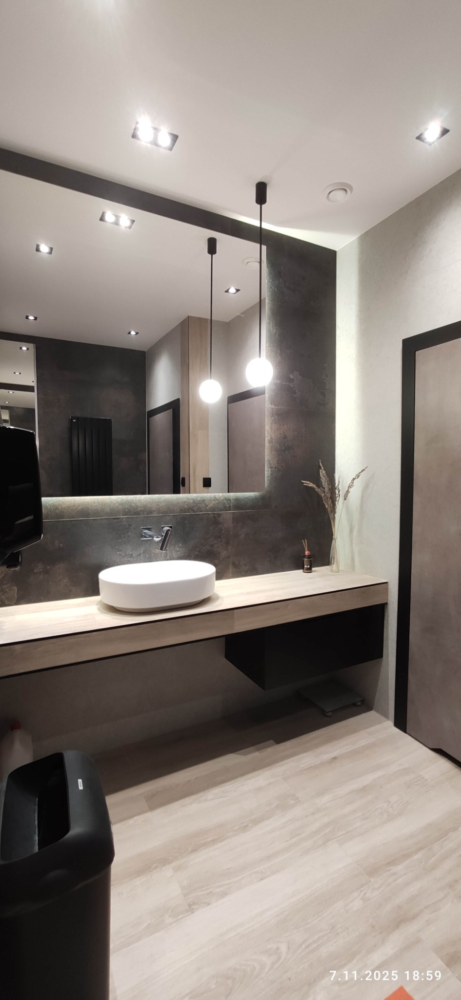 Nowoczesna łazienka z owalną umywalką nablatową, podświetlanym lustrem i wiszącymi lampami kulami. Minimalistyczny design z elementami drewna i szarości.