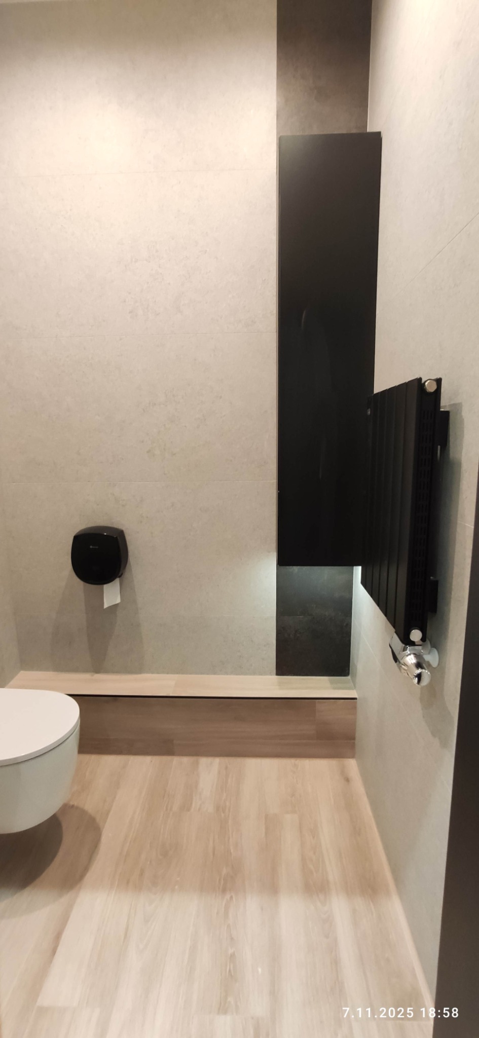 Wykończona łazienka z beżowymi płytkami, czarnym grzejnikiem i szafką. Jasna podłoga imitująca drewno. Widoczny fragment toalety i dozownik papieru.
