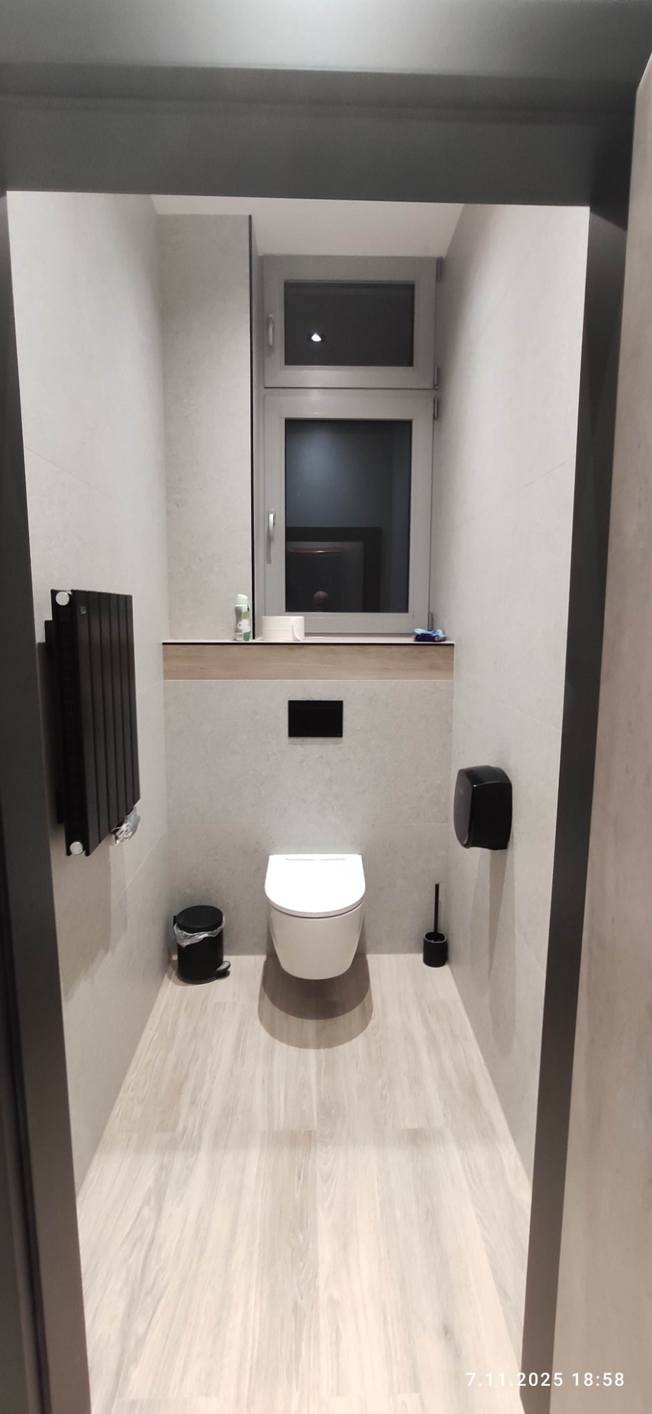 Wąska, jasna toaleta z podwieszaną muszlą, grzejnikiem drabinkowym i drewnopodobną podłogą. Nad muszlą okno z lustrem i półką. Minimalistyczny design.