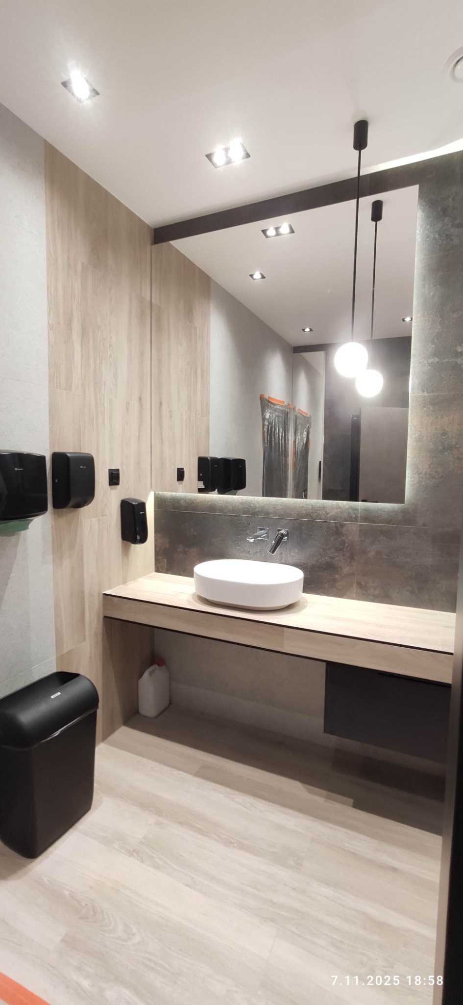 Nowoczesna łazienka publiczna z okrągłą umywalką, podświetlanym lustrem i drewnopodobnymi płytkami. Widoczne dozowniki mydła i kosz na śmieci.