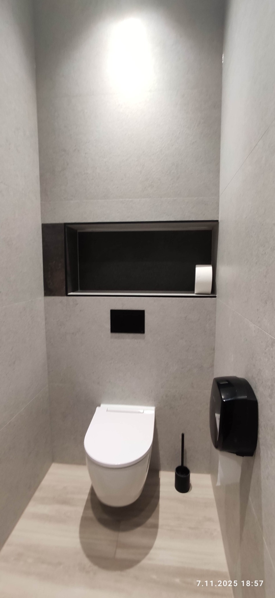 Nowoczesna łazienka z wiszącą toaletą, szarą glazurą i wnęką z rolką papieru. Minimalistyczny design, drewnopodobna podłoga, czarne akcenty.