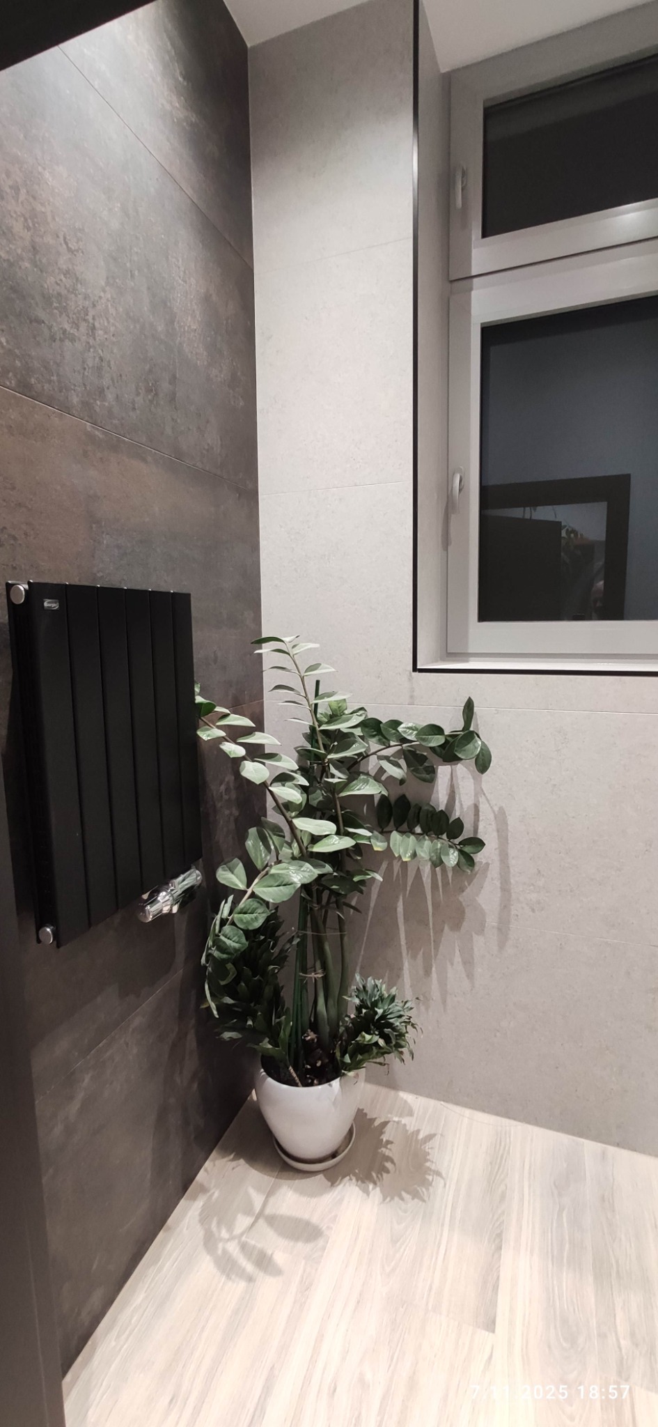 Nowoczesna łazienka z szarymi płytkami, czarnym grzejnikiem i rośliną w białej doniczce. Minimalistyczny design, jasne drewnopodobne panele podłogowe.