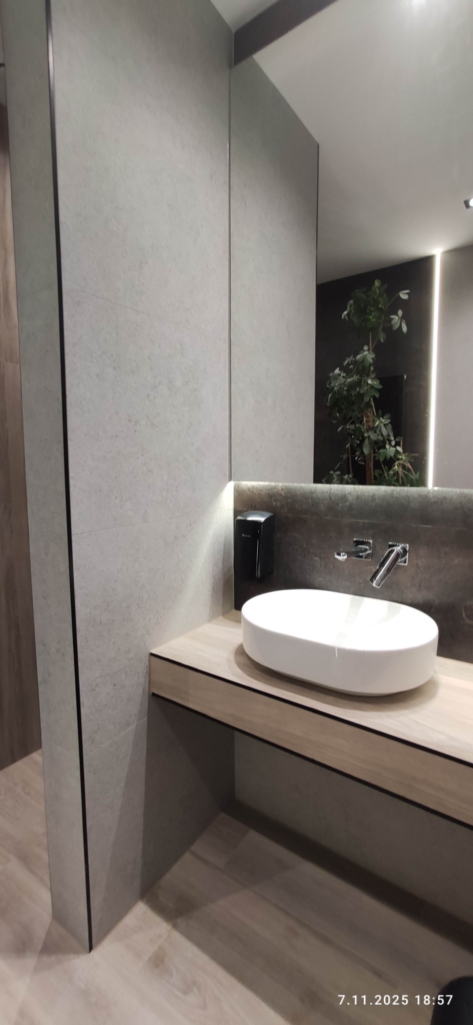 Nowoczesna łazienka z owalną umywalką nablatową, szarą płytką imitującą kamień i minimalistyczną armaturą. W tle lustro z odbiciem rośliny doniczkowej.