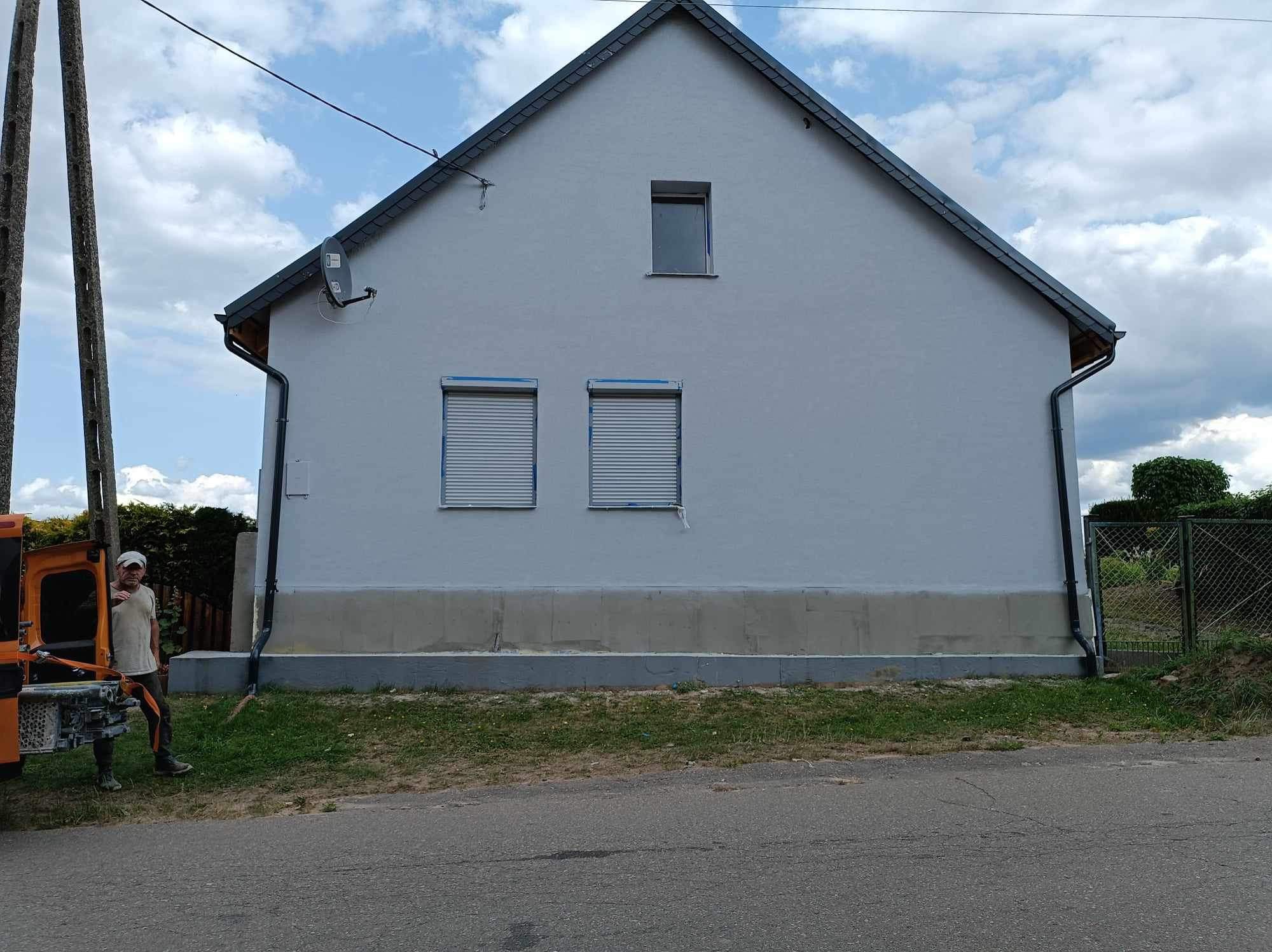Świeżo pomalowana elewacja domu jednorodzinnego w odcieniach szarości, z oknami zabezpieczonymi taśmą malarską, widoczny fragment samochodu dostawczego i pracownik w tle.