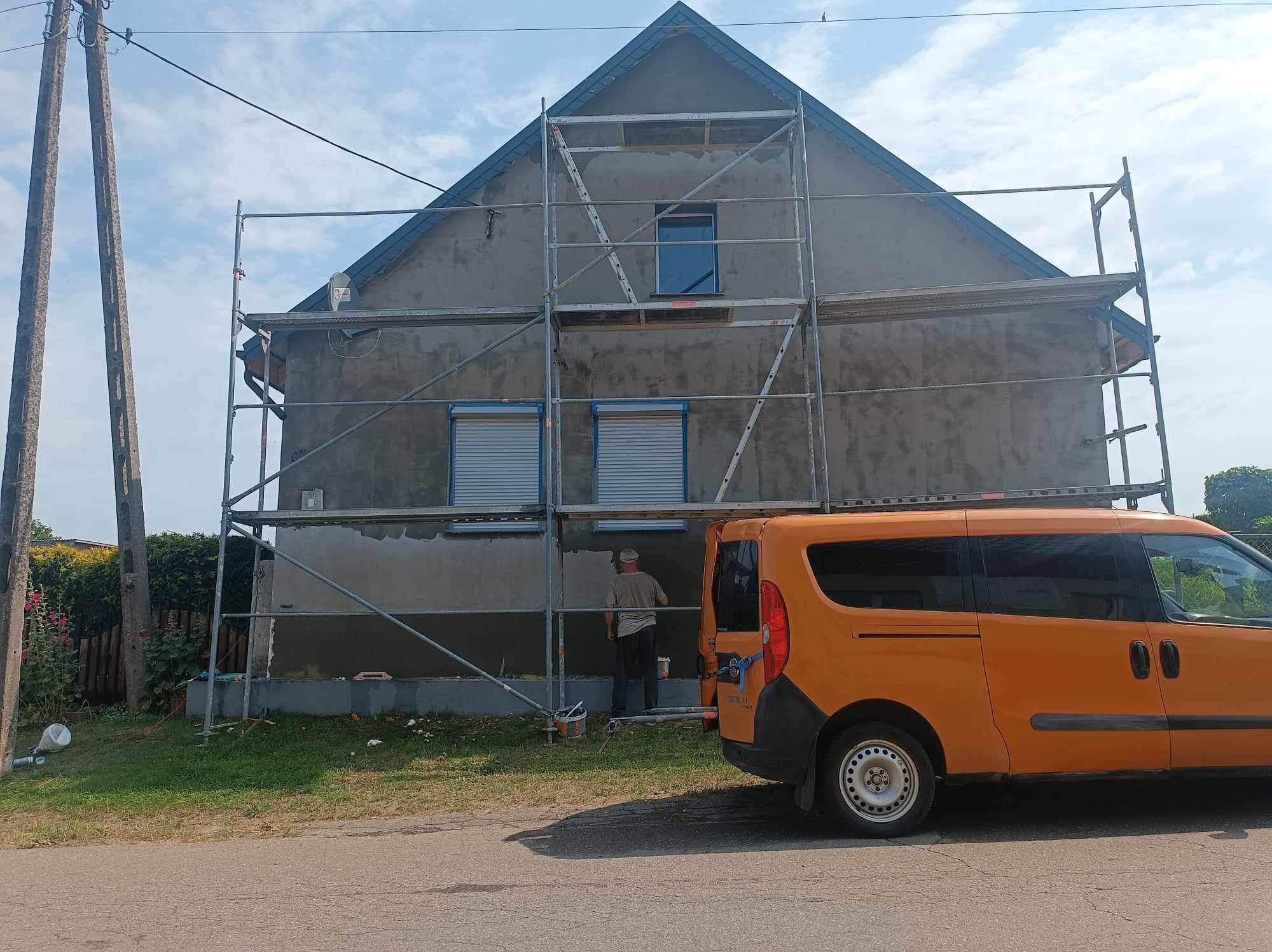 Pracownik w trakcie nakładania szarej warstwy tynku na elewację domu jednorodzinnego, widoczne rusztowanie i pomarańczowy samochód dostawczy.