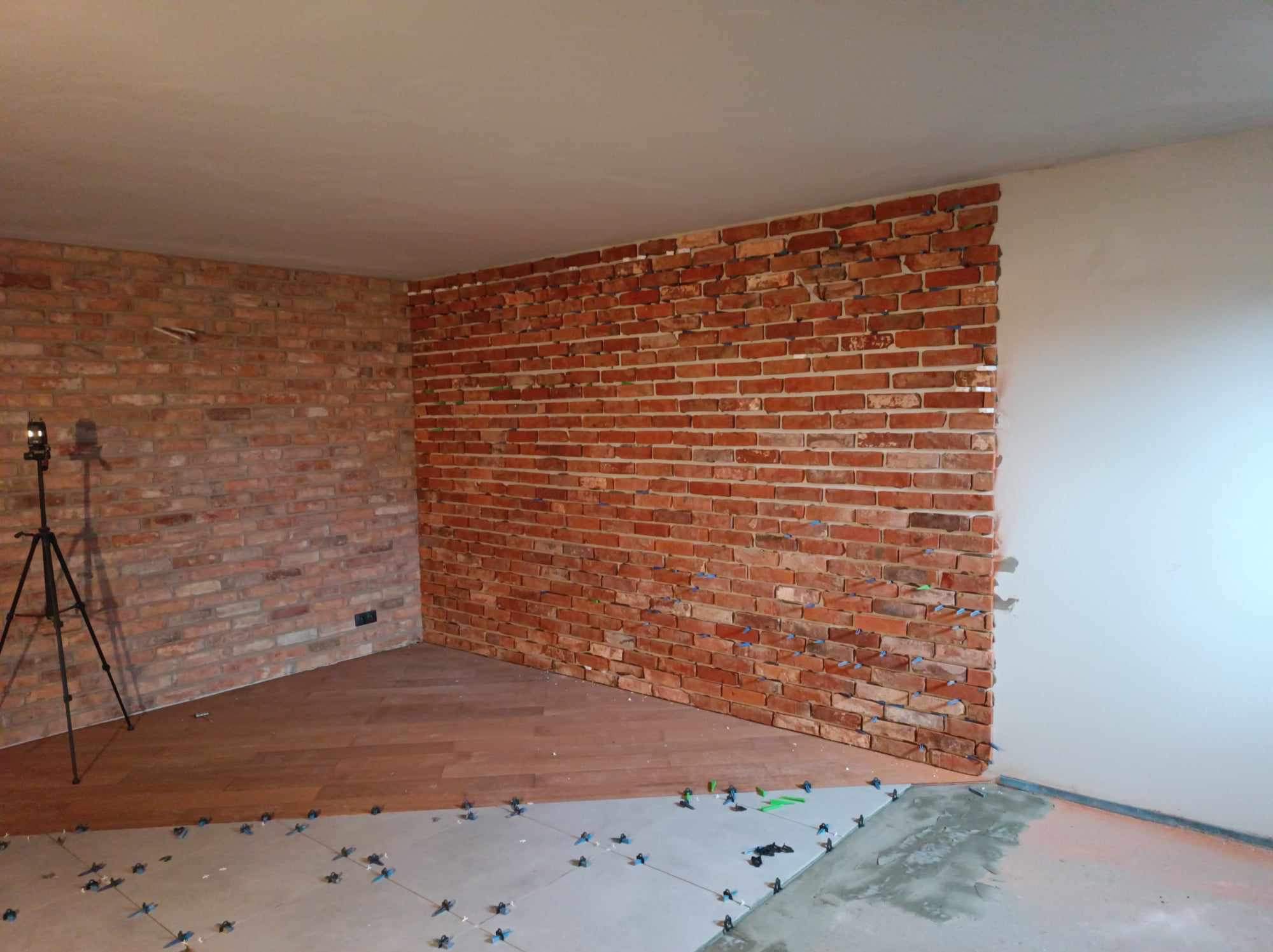 Wnętrze w trakcie wykańczania: fragment ściany z cegły klinkierowej z niebieskimi krzyżykami dystansowymi, częściowo położona drewniana podłoga, widoczny statyw aparatu.