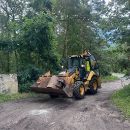 GAPKOP PATRYK GAPSKI - Żółta koparko-ładowarka Caterpillar na ubitej drodze gruntowej, otoczona drzewami, operator w kamizelce odblaskowej w kabinie, widoczny betonowy element z graffiti w tle.