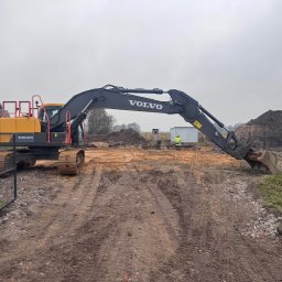 GAPKOP PATRYK GAPSKI - Żółto-czarna koparka Volvo na placu budowy z otwartą bramą, widoczny operator w odblaskowej kamizelce i pryzma ziemi w tle, pochmurne niebo.