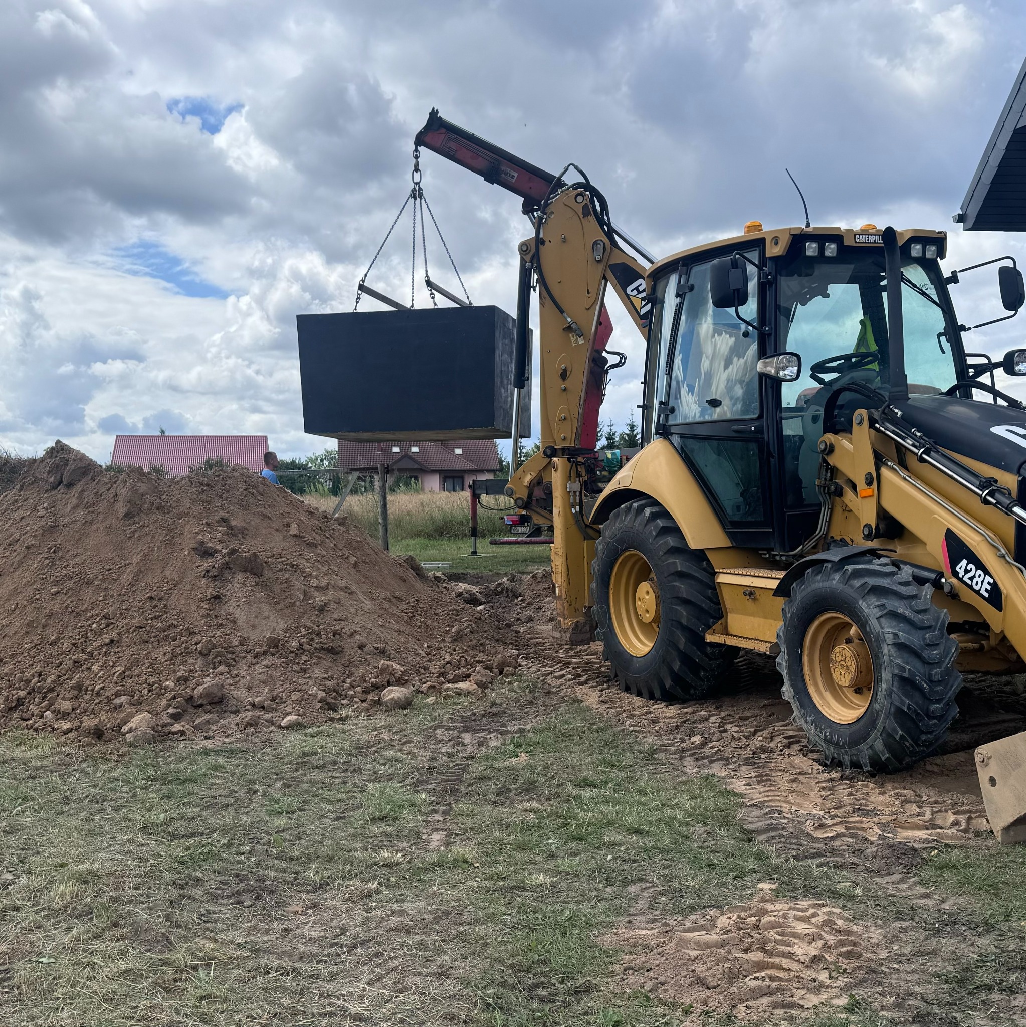Żółta koparko-ładowarka Caterpillar 428E podnosi czarny, betonowy zbiornik nad wykopem na tle pochmurnego nieba i domów z czerwonymi dachami.