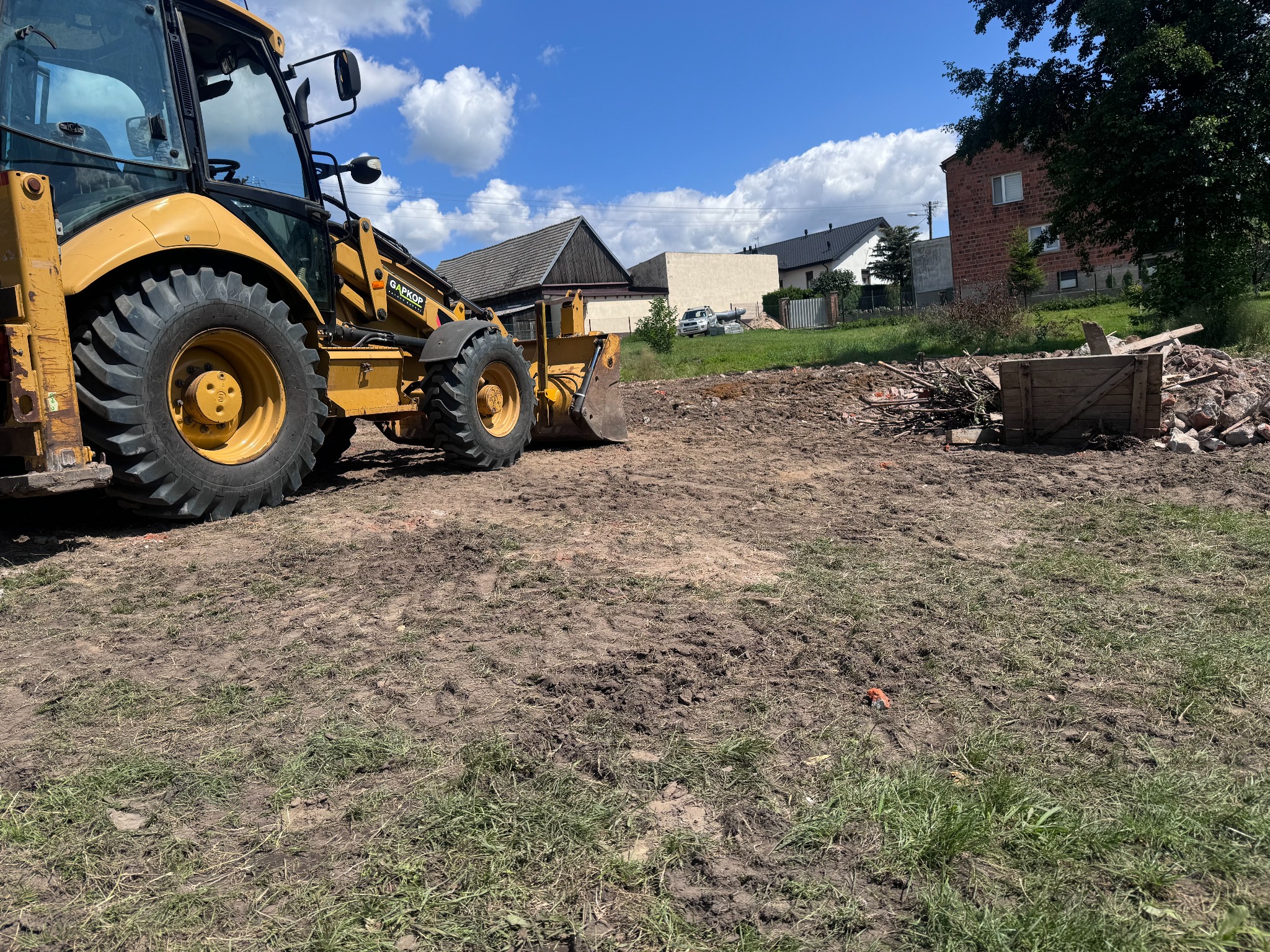 Żółta koparko-ładowarka na nierównym, błotnistym terenie z gruzem i drewnianą skrzynią w tle, domy i błękitne niebo z chmurami w oddali.