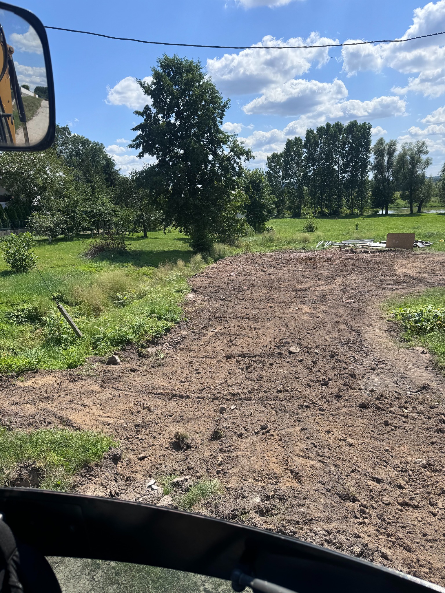 Wyrównany teren z widocznymi śladami opon, zielona trawa po bokach, w tle drzewa i błękitne niebo z chmurami, widok z kabiny maszyny budowlanej.