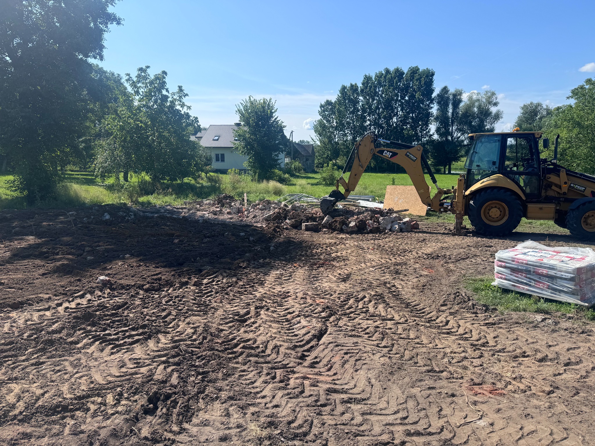Żółta koparko-ładowarka CAT na placu budowy, w tle gruz i fragmenty rozbiórki, widoczne ślady opon na ziemi i paleta z materiałami budowlanymi. Krajobraz wiejski z domem i drzewami w tle.