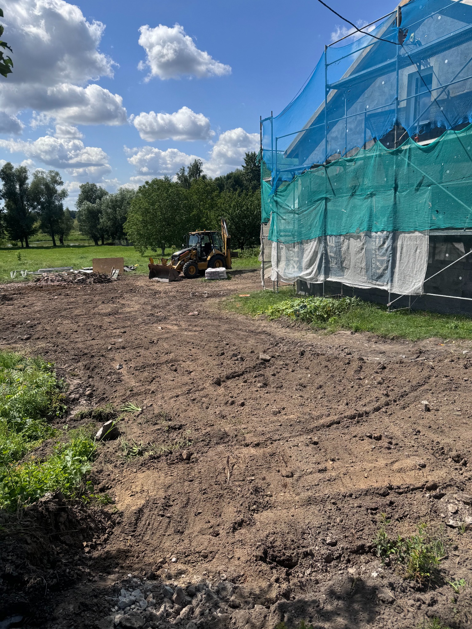 Żółta koparko-ładowarka na placu budowy z częściowo rozebranym budynkiem zabezpieczonym siatką, widoczne ślady opon w ziemi i fragmenty gruzu.