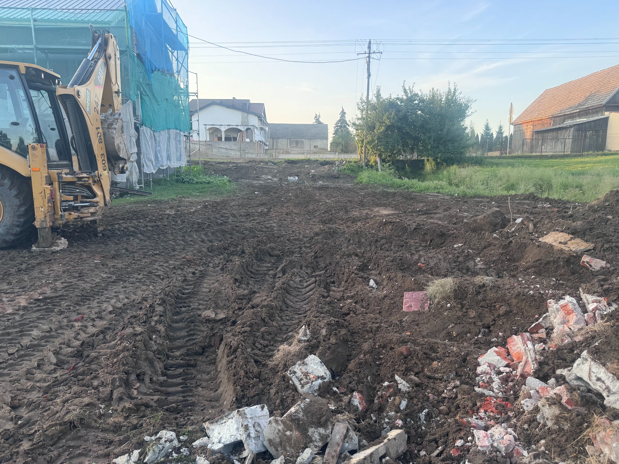 Żółta koparko-ładowarka CAT na placu budowy po wyburzeniu, gruz i ślady opon na ubitej ziemi, w tle budynki mieszkalne i rusztowania zabezpieczające.