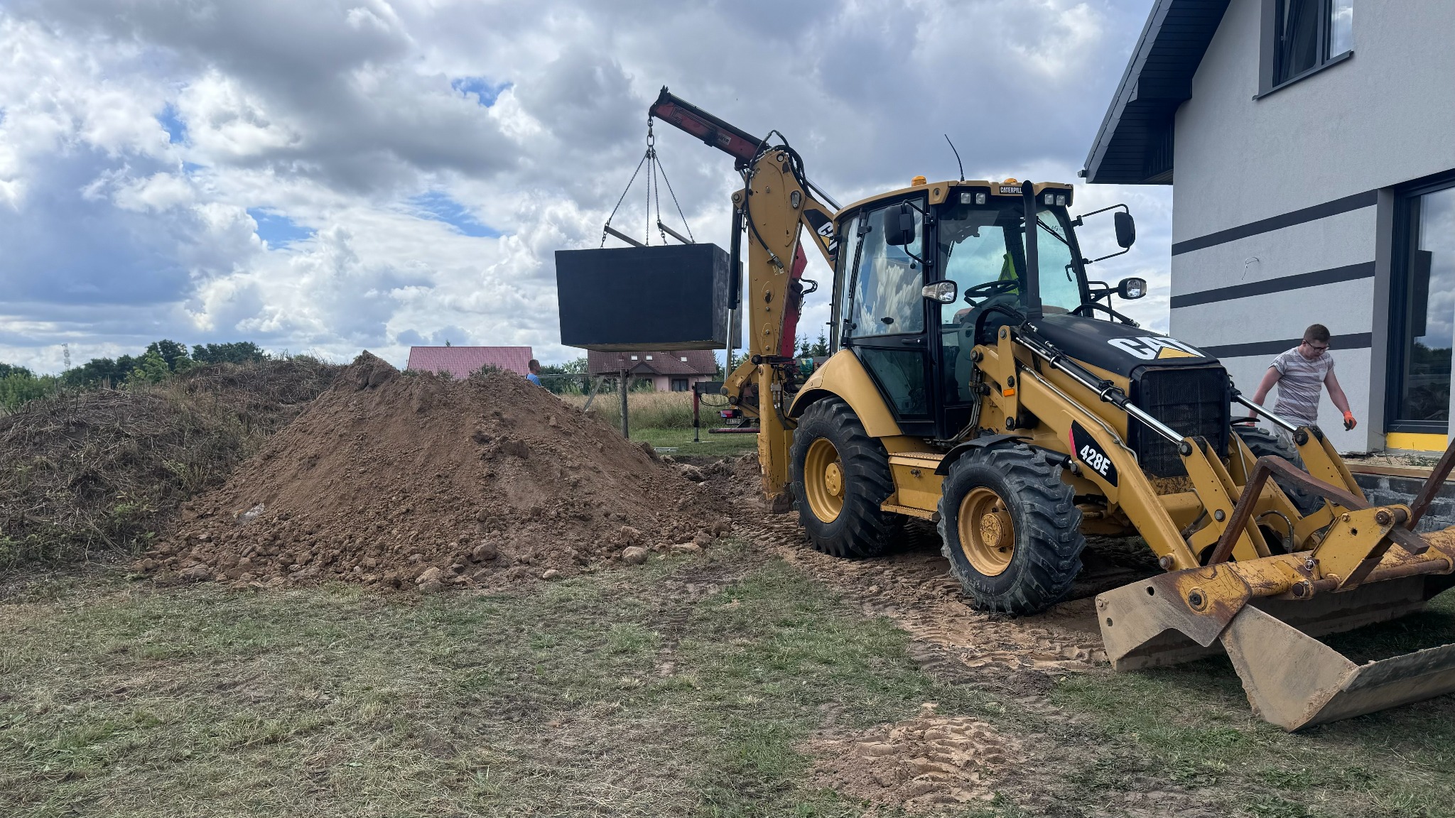 Żółta koparko-ładowarka Caterpillar 428E podnosi za pomocą łańcuchów betonowy zbiornik na tle nowo wybudowanego domu z szarą elewacją i stertą ziemi.