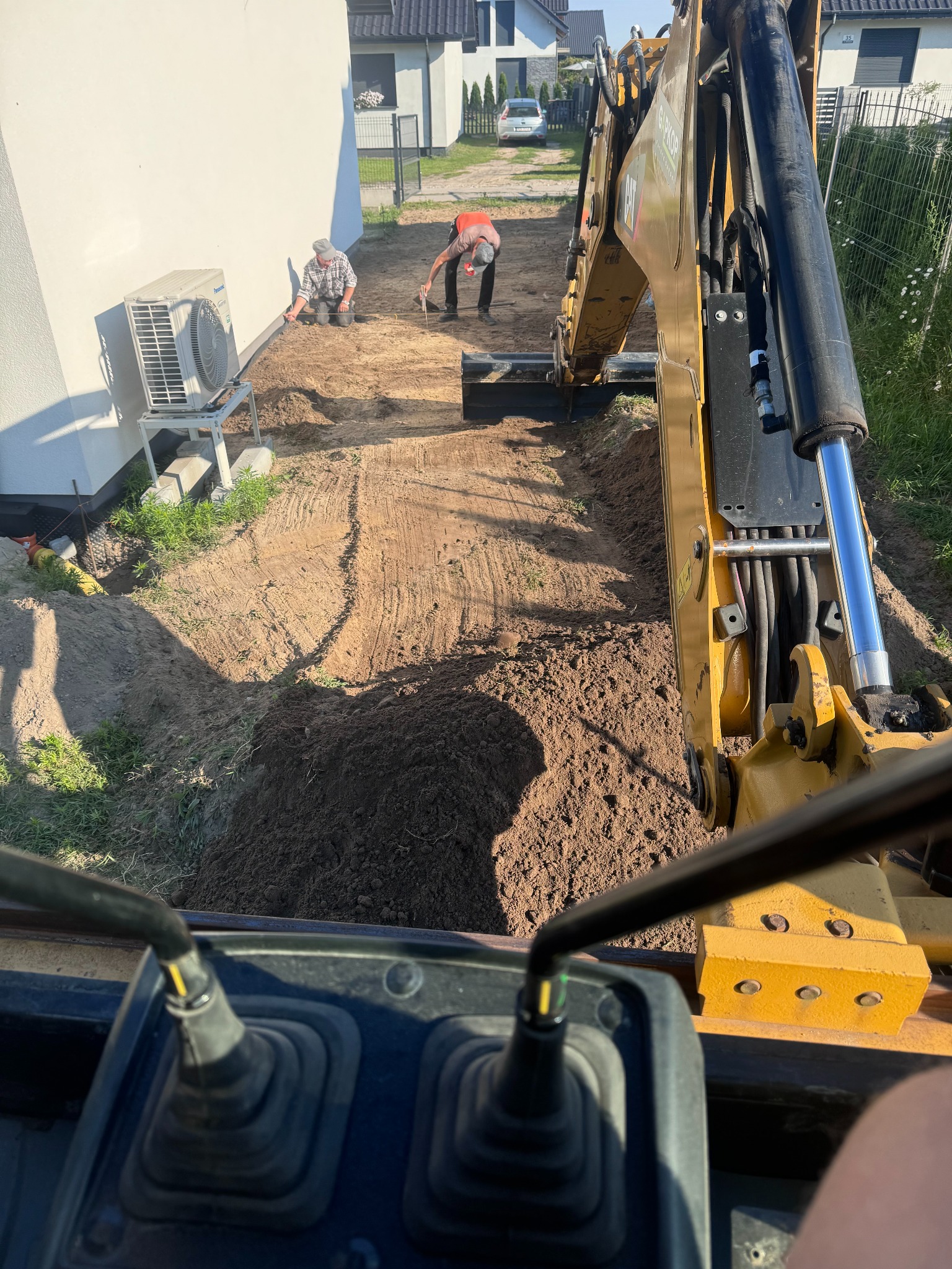 Żółta koparka w trakcie prac ziemnych na działce, widok z kabiny operatora, dwóch pracowników w oddali przygotowuje teren wzdłuż fundamentów domu.