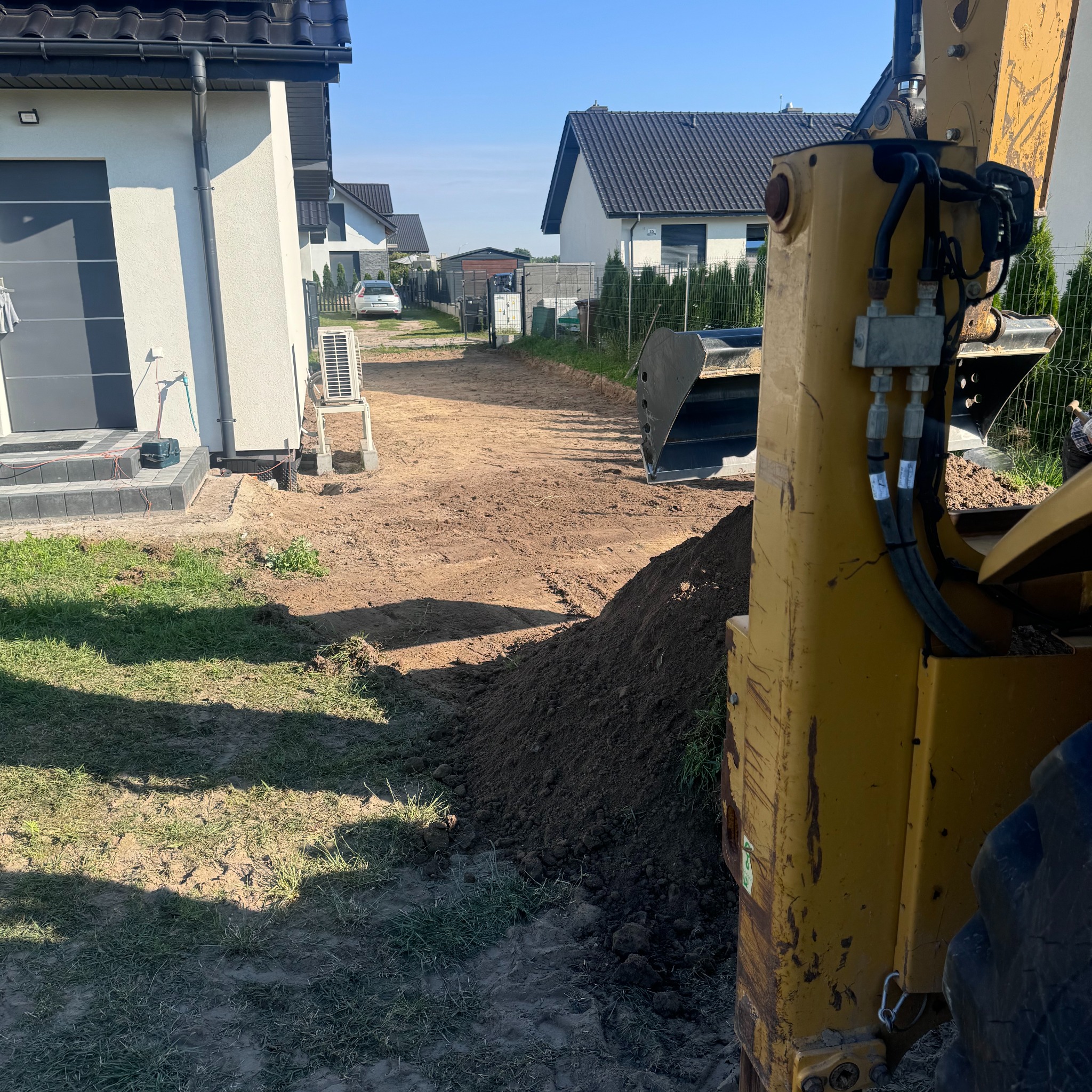 Żółta koparka częściowo widoczna na pierwszym planie, w tle wyrównany teren działki budowlanej z widocznymi domami jednorodzinnymi w słoneczny dzień.