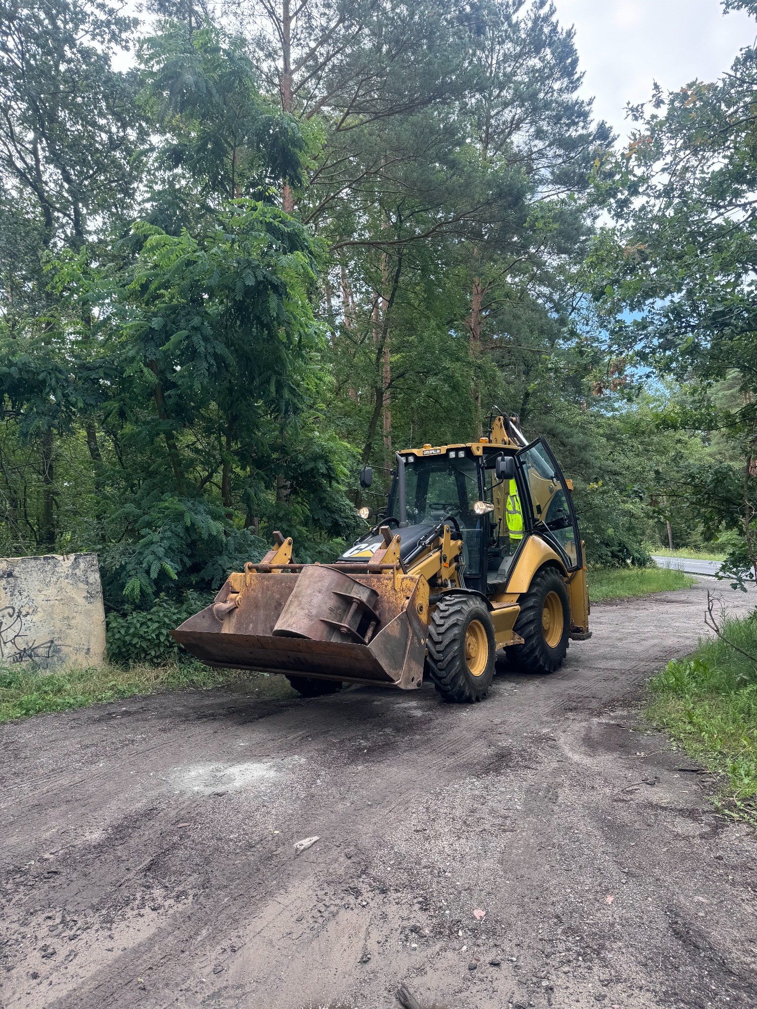 Żółta koparko-ładowarka Caterpillar na ubitej drodze gruntowej, otoczona drzewami, operator w kamizelce odblaskowej w kabinie, widoczny betonowy element z graffiti w tle.