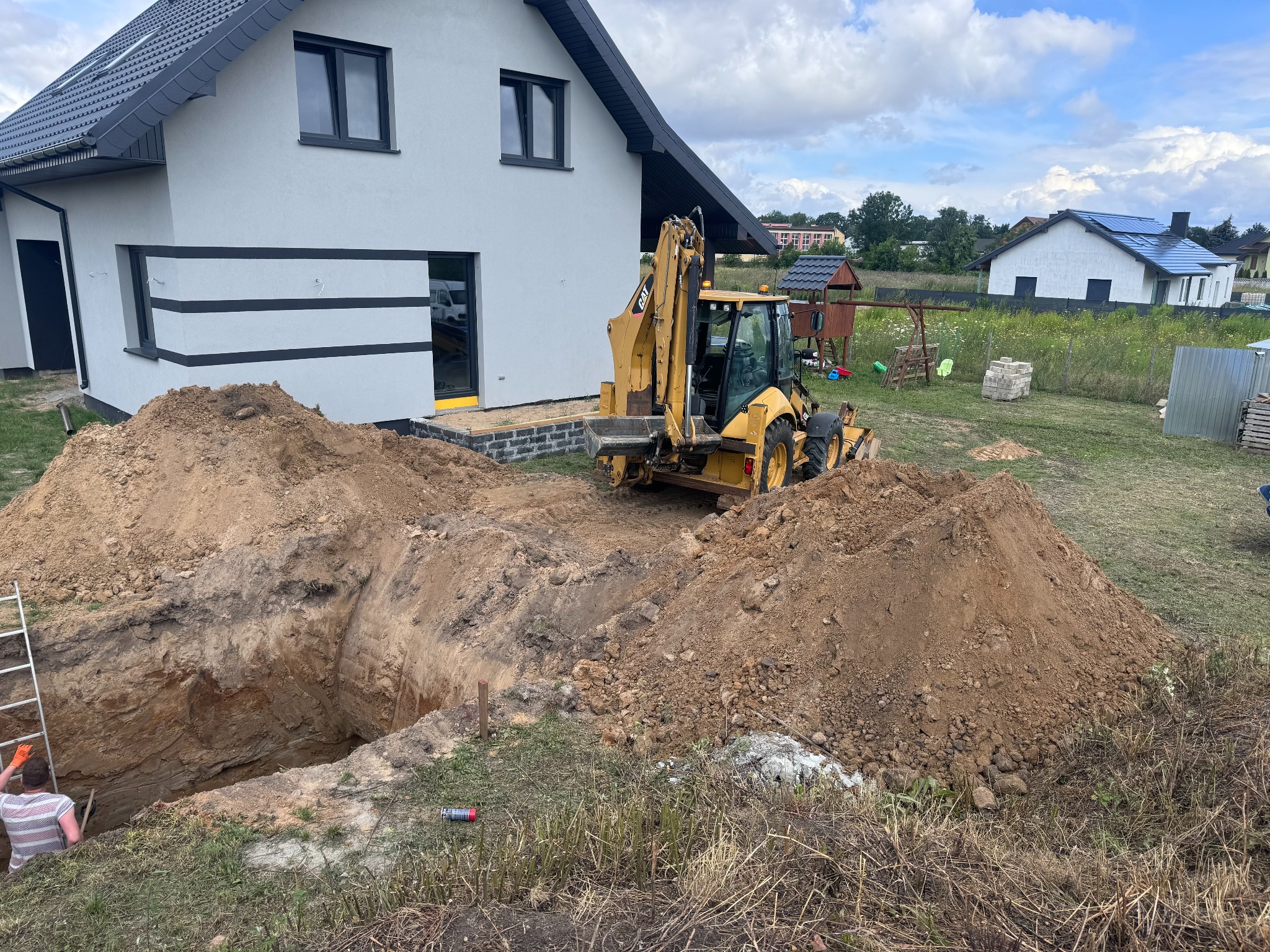 Żółta koparko-ładowarka CAT przy wykopie w ziemi obok nowego domu z szarą elewacją i czarnymi akcentami. W tle widoczny ogród i inne budynki.