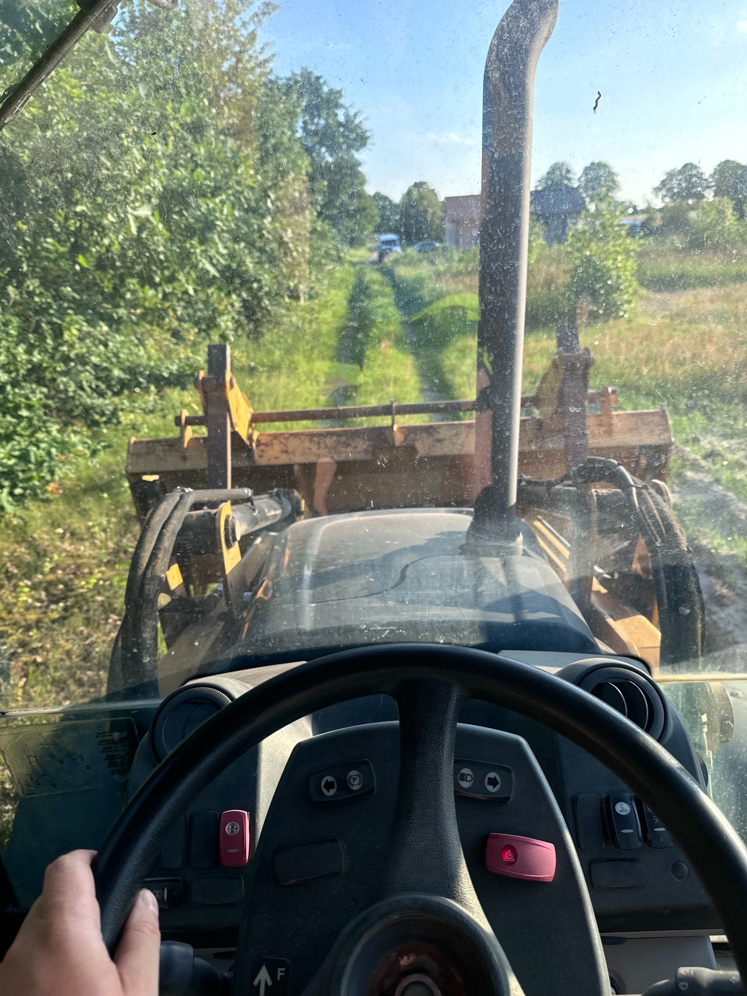 Widok z kabiny operatora na maszynę budowlaną (ładowarka teleskopowa?) w trakcie pracy na zewnątrz, z widocznym fragmentem kierownicy i dłoni operatora, oraz rurą wydechową na tle zieleni i nieba.