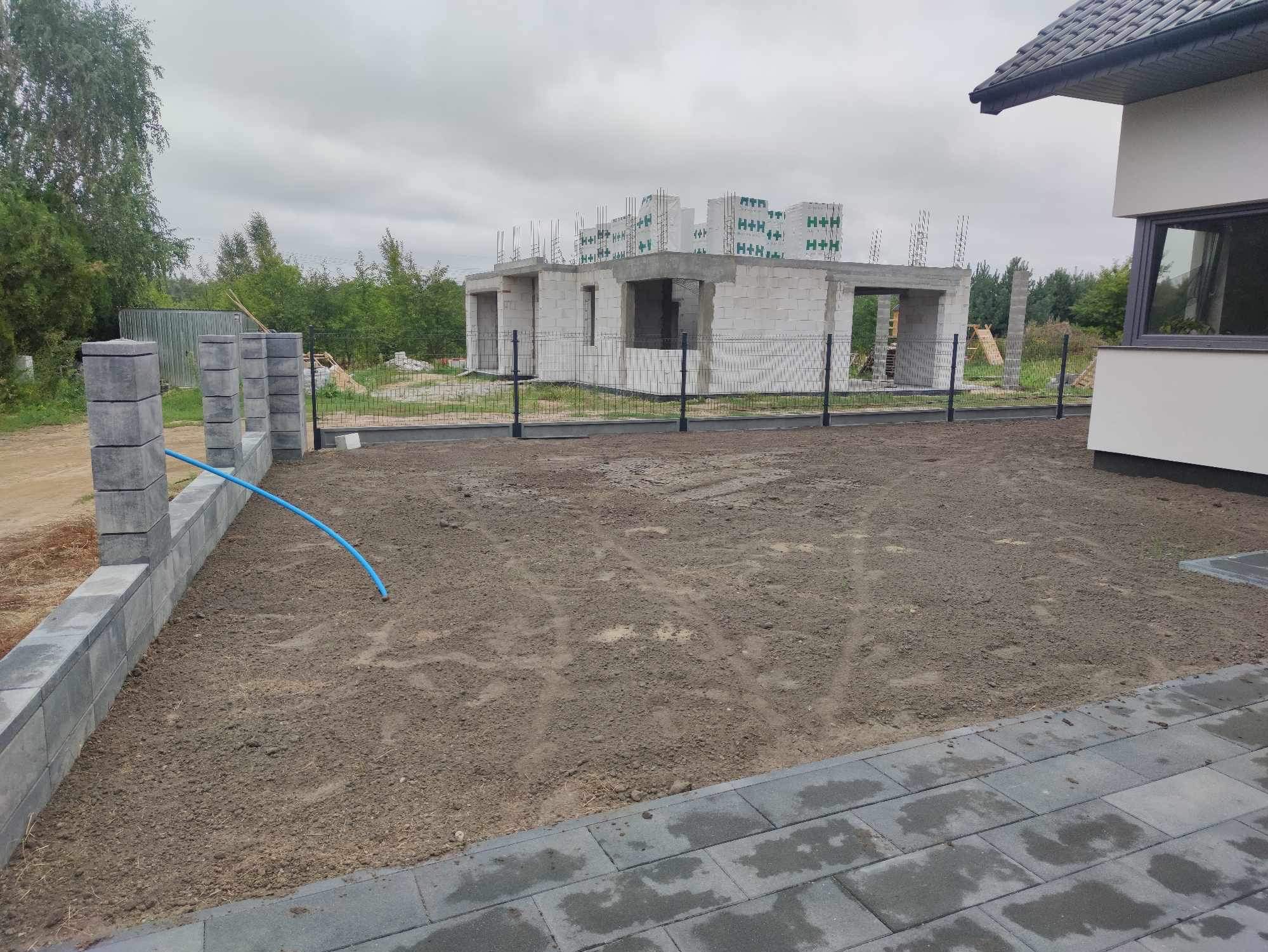 Surowy stan budowy domu z bloczków betonowych, widok z poziomu gruntu z nowo postawionym, szarym ogrodzeniem z betonowych słupków i podmurówki, z widoczną niebieską rurą nawadniającą.