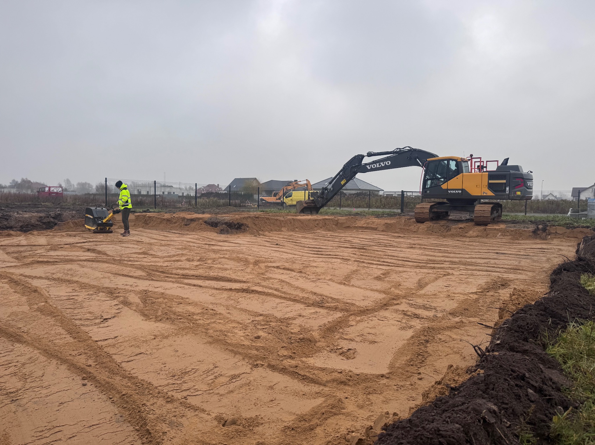 Przygotowanie terenu pod budowę: wyrównana piaskowa powierzchnia z widocznymi śladami maszyn, operator zagęszczarki w żółtej kamizelce, w tle koparka Volvo i zabudowa jednorodzinna w pochmurny dzień.