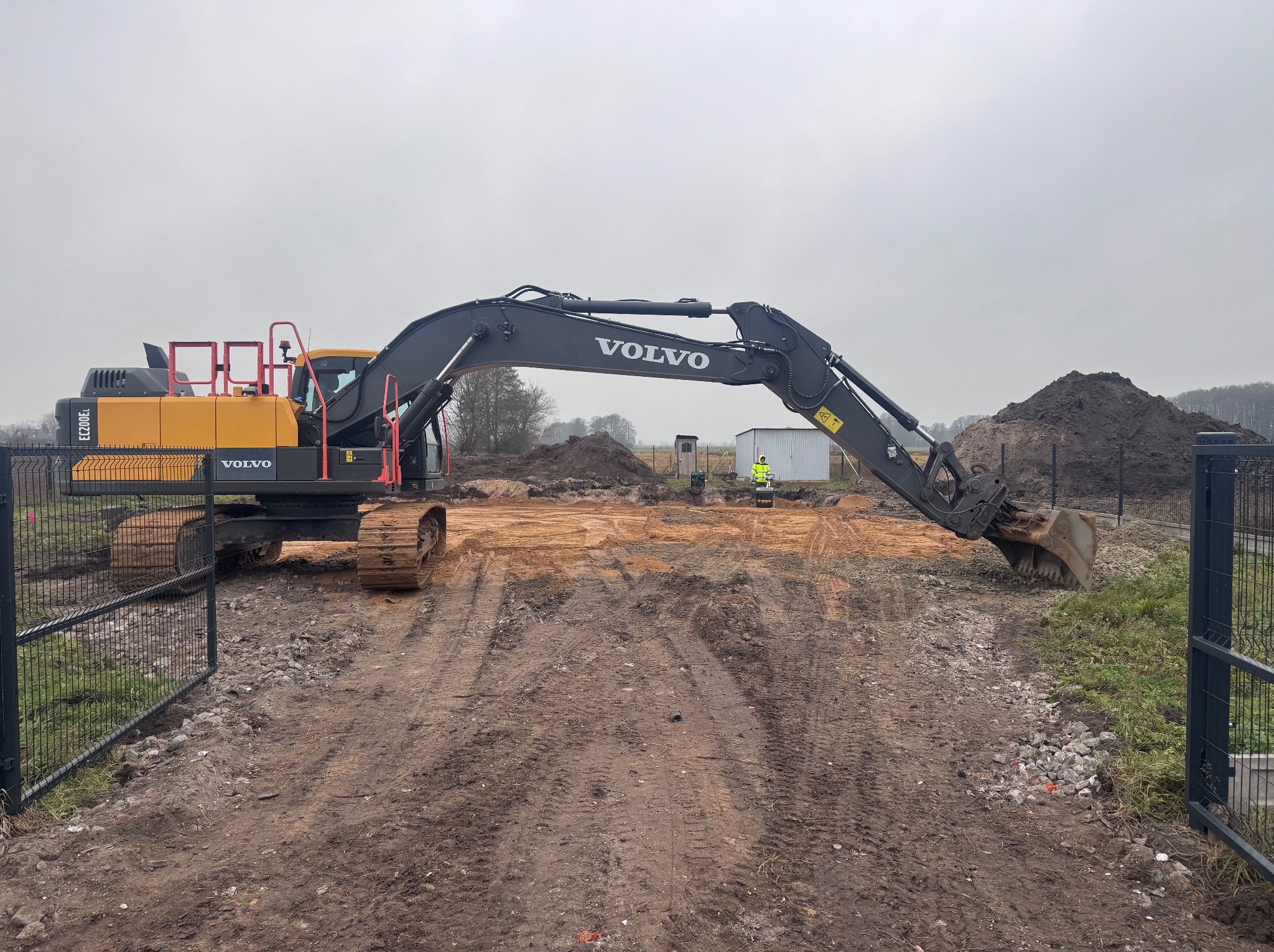 Żółto-czarna koparka Volvo na placu budowy z otwartą bramą, widoczny operator w odblaskowej kamizelce i pryzma ziemi w tle, pochmurne niebo.