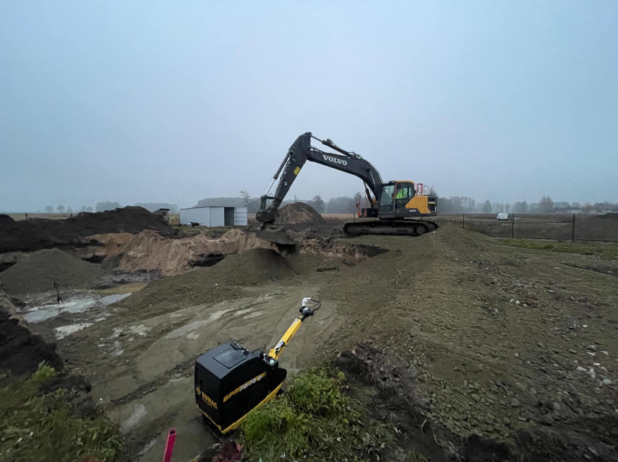 Żółto-czarna koparka Volvo na placu budowy, w tle sterta ziemi i budynek gospodarczy, na pierwszym planie zagęszczarka gruntu Bomag BPR70/70D.