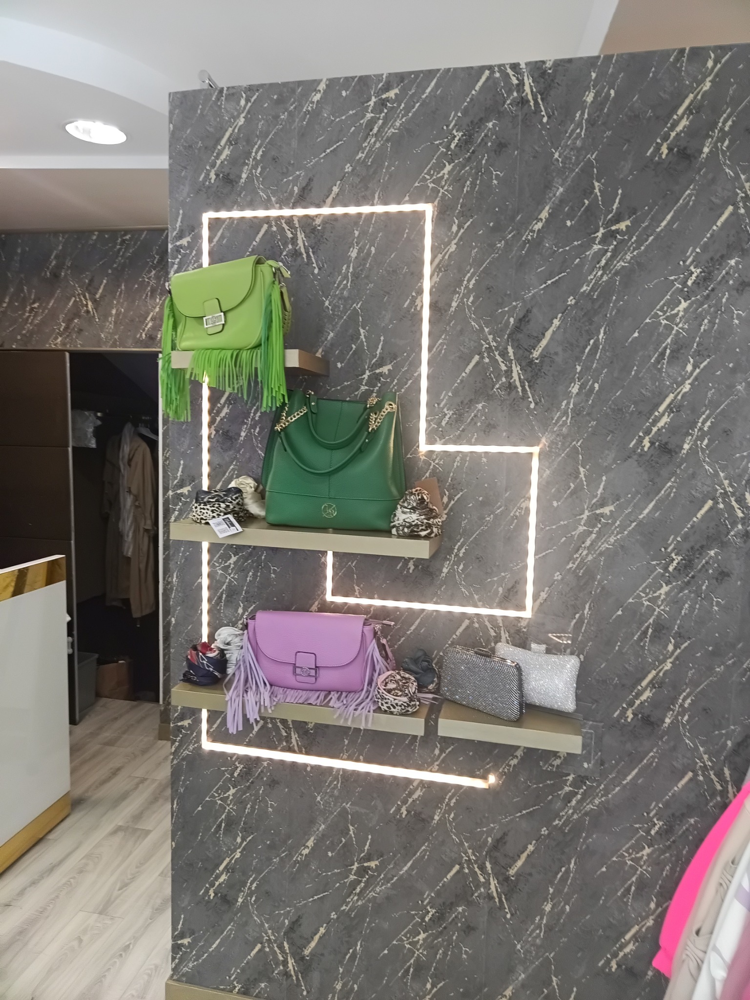 Wnętrze butiku z szarą ścianą imitującą marmur, oświetloną ledowymi kwadratami, na której zamontowano półki z torebkami w odcieniach zieleni i fioletu, oraz dodatkami.