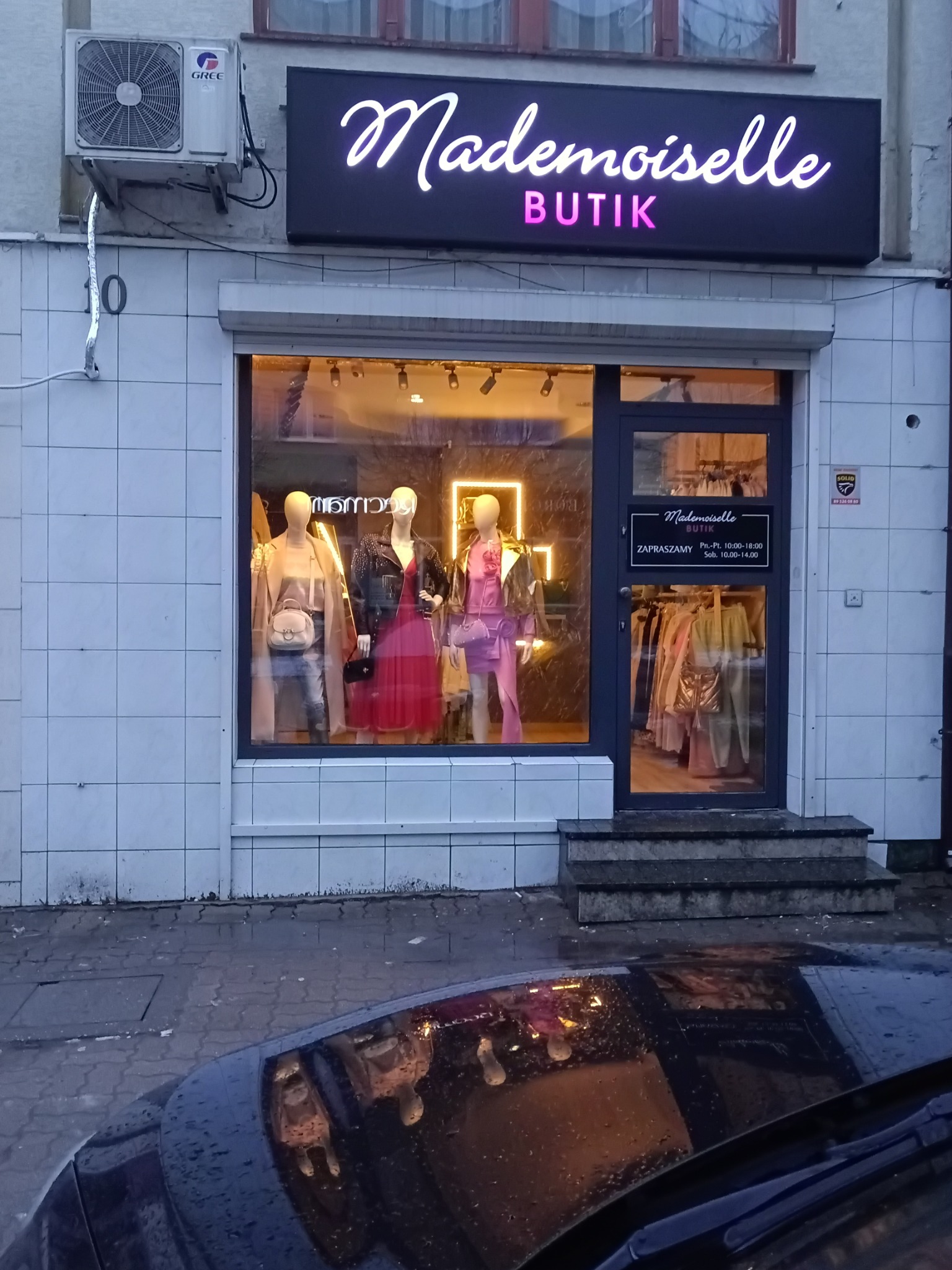 Fasada butiku Mademoiselle z neonowym szyldem, manekiny w oknie wystawowym prezentujące modne ubrania, odbicie w mokrej karoserii samochodu na pierwszym planie.