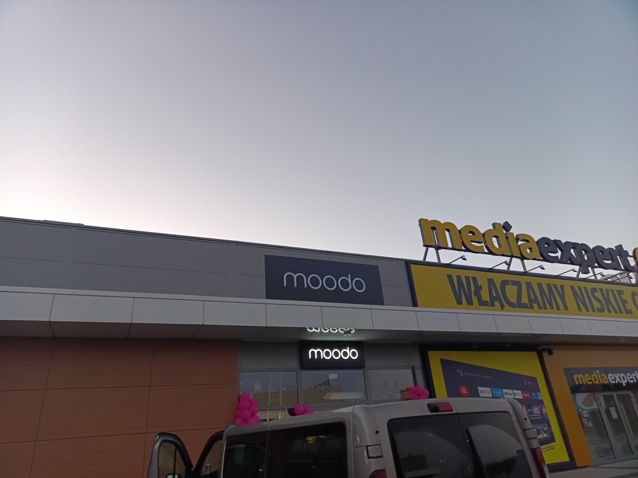 Elewacja budynku z szyldami 'moodo' i 'mediaexpert', częściowo zasłonięta przez samochód dostawczy, z dekoracjami balonowymi sugerującymi otwarcie lub promocję.