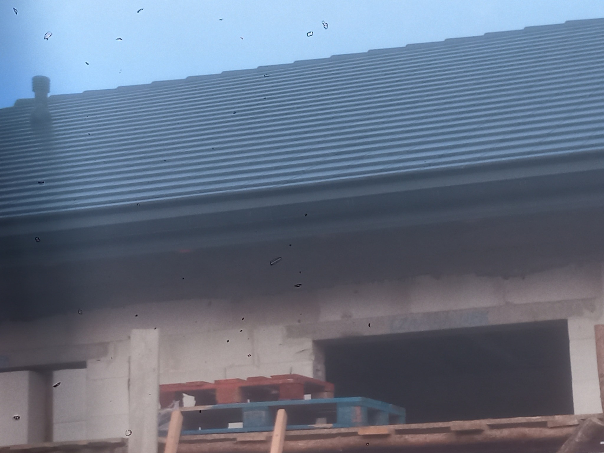 Szary dach z blachodachówki z kominem, widoczny fragment surowego stanu domu w budowie z paletami wewnątrz.