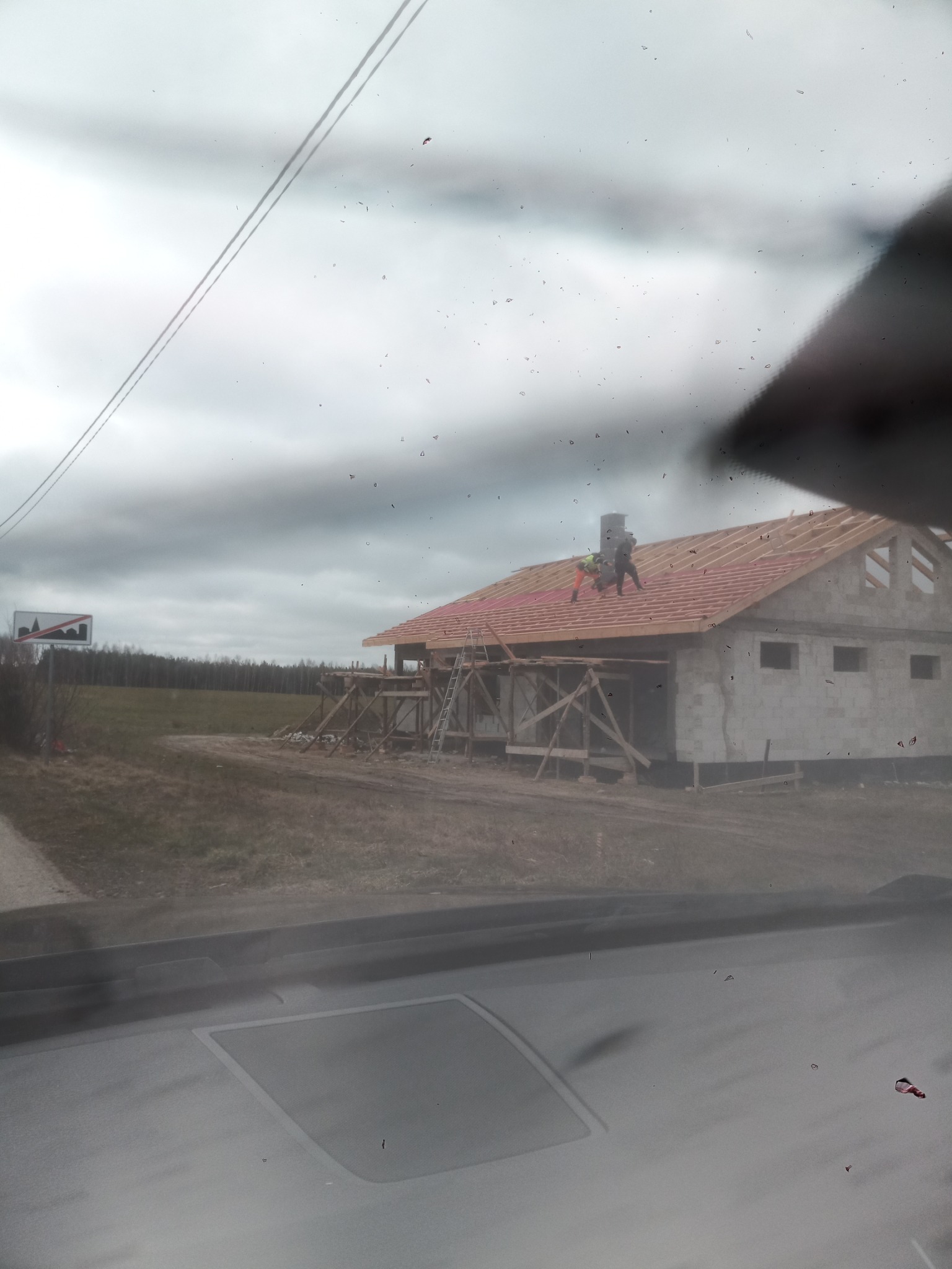 Budowa domu w trakcie realizacji: murarz na dachu układa czerwoną dachówkę, widoczne rusztowania, surowy stan budynku z bloczków, pochmurne niebo w tle, zdjęcie zrobione zza szyby samochodu.