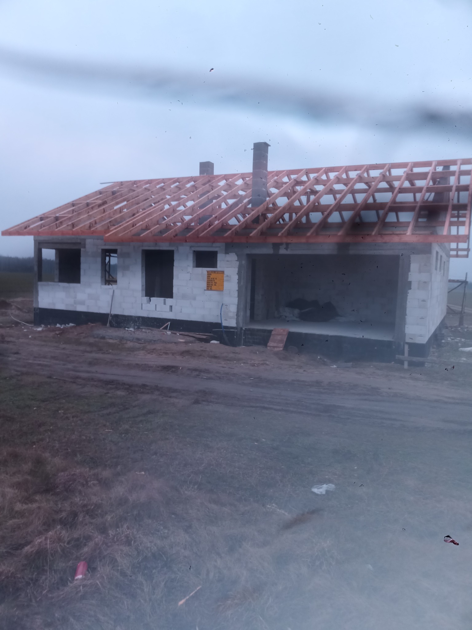 Surowy stan domu murowanego z bloczków betonowych, widoczny szkielet dachu z drewnianych krokwi, komin z cegły, teren budowy z ziemią i trawą, pochmurne niebo w tle.