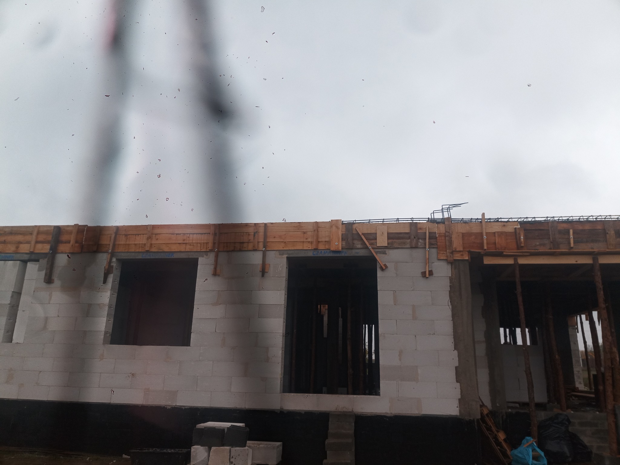 Ściana domu murowanego w trakcie budowy, z widocznymi otworami okiennymi i drewnianymi szalunkami na stropie, oraz bloczkami betonu komórkowego u podstawy.