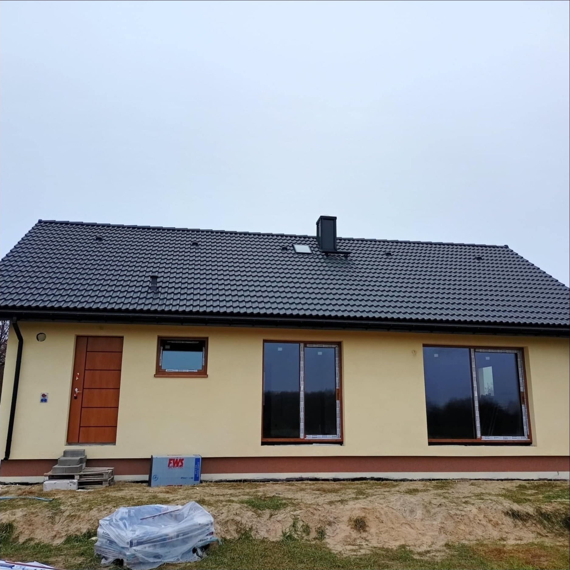 Nowo wybudowany dom jednorodzinny z ciemną dachówką, beżową elewacją i brązowymi oknami, widoczne materiały budowlane na trawniku przed domem.