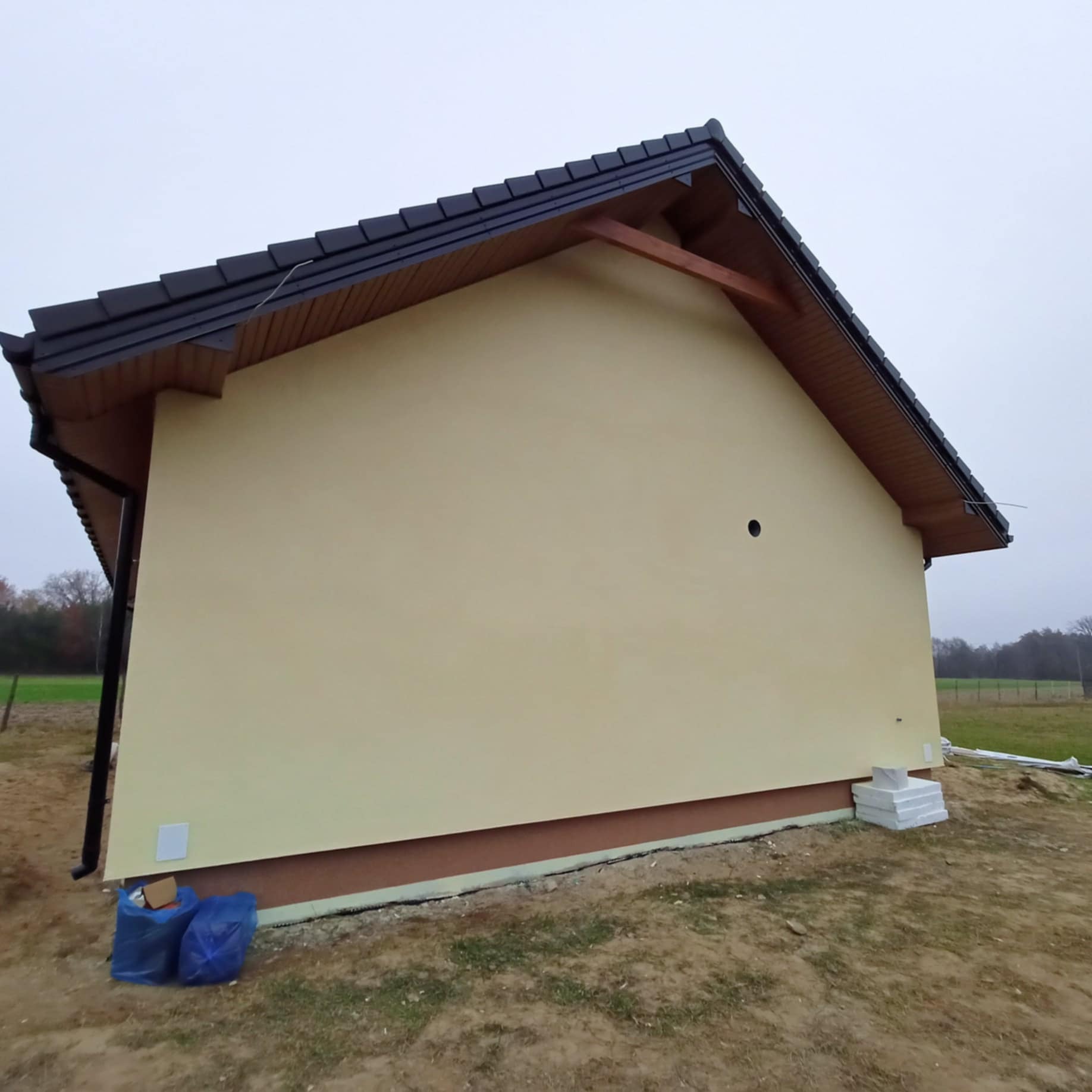 Świeżo otynkowany, beżowy budynek z ciemną dachówką i widocznym fragmentem ocieplenia fundamentu, z niebieskimi workami i styropianem leżącymi obok.
